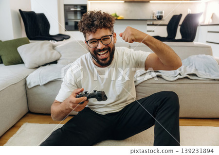 Celebrating victory, playing video game by using controller. Man is in the modern domestic room at home 139231894