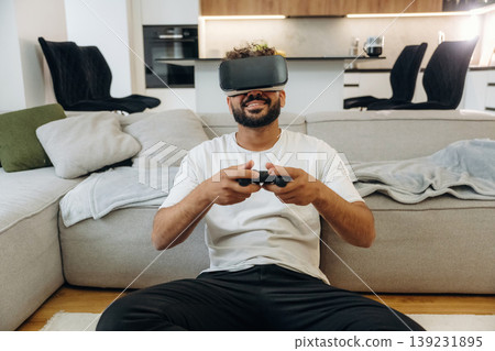 Virtual reality glasses, controller in hands. Man is in the modern domestic room at home Virtual reality glasses, controller in hands. Man is in the modern domestic room at home 139231895