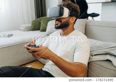 Virtual reality glasses, controller in hands. Man is in the modern domestic room at home Virtual reality glasses, controller in hands. Man is in the modern domestic room at home 139231896