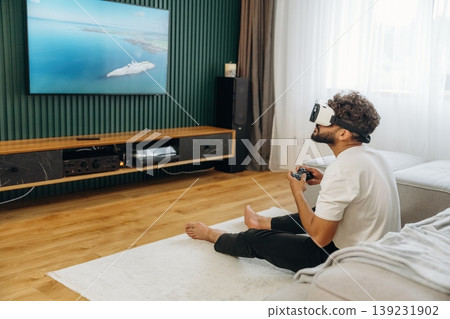 On the floor, in virtual reality headset, holding controller. Man is in the modern domestic room at home On the floor, in virtual reality headset, holding controller. Man is in the modern domestic room at home 139231902