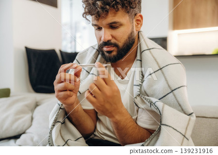 Body temperature measuring by using thermometer. Sick man is on the sofa at home, with towel, feeling bad Body temperature measuring by using thermometer. Sick man is on the sofa at home, with towel, feeling bad 139231916