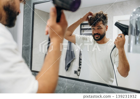 Using hair dryer. Guy is in the bathroom, taking care of his appearance Using hair dryer. Guy is in the bathroom, taking care of his appearance 139231932