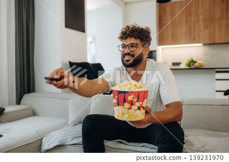 Popcorn and TV controller. Man is in the modern domestic room at home Popcorn and TV controller. Man is in the modern domestic room at home 139231970