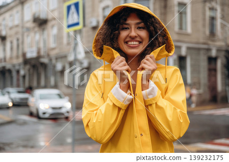 In yellow raincoat, standing and smiling. Happy young woman in the city during the rain In yellow raincoat, standing and smiling. Happy young woman in the city during the rain 139232175