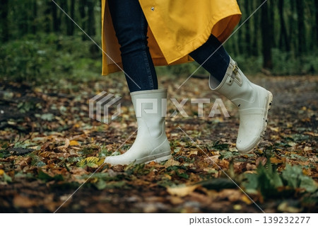 Having a walk. Close up view of woman legs, in rainy forest, yellow raincoat and rubber boots Having a walk. Close up view of woman legs, in rainy forest, yellow raincoat and rubber boots 139232277