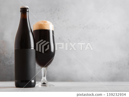 Bottle and glass of fresh cold dark stout beer with foam on light table background. Bottle and glass of fresh cold dark stout beer with foam on light table background. 139232934