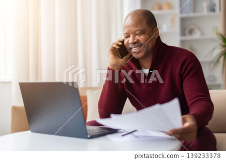 Smiling mature black man talking on smartphone and reviewing documents at home Smiling mature black man talking on smartphone and reviewing documents at home 139233378