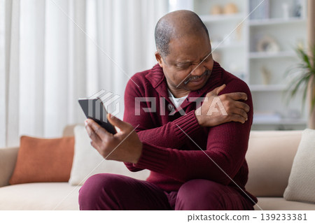 Mature black man grimacing in shoulder pain while holding smartphone at home Mature black man grimacing in shoulder pain while holding smartphone at home 139233381