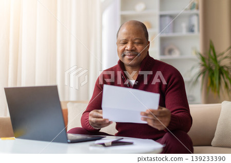 Pleased mature African American man reading paper document beside laptop at home Pleased mature African American man reading paper document beside laptop at home 139233390