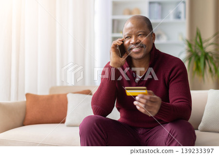 Mature black man talking on phone while holding credit card at home Mature black man talking on phone while holding credit card at home 139233397