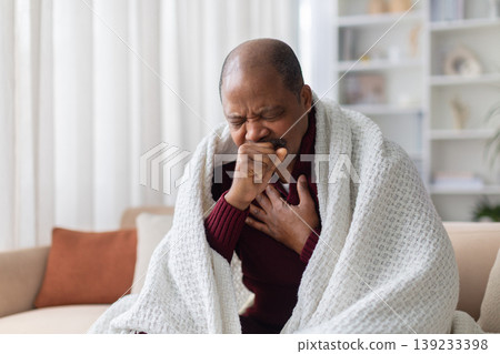 Sick mature African American man coughing into fist on sofa at home Sick mature African American man coughing into fist on sofa at home 139233398