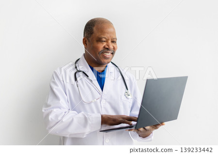 Happy mature African American male doctor in white lab coat using laptop Happy mature African American male doctor in white lab coat using laptop 139233442