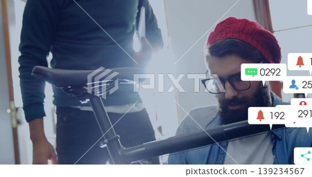 Man wearing cap working on bicycle frame, saddle in studio, with tablet, social notification icons Man wearing cap working on bicycle frame, saddle in studio, with tablet, social notification icons 139234567