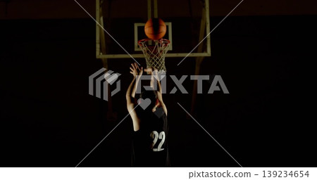 Reaching up athlete wearing number 22 jersey shooting basketball in dark gym, with lit hoop, net Reaching up athlete wearing number 22 jersey shooting basketball in dark gym, with lit hoop, net 139234654