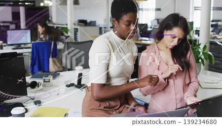 Collaborating coworkers wearing cream top and light pink blouse pointing at laptop in modern office Collaborating coworkers wearing cream top and light pink blouse pointing at laptop in modern office 139234680