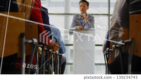 Speaking presenter wearing grey blazer at conference front holding mic with audience taking notes 139234743