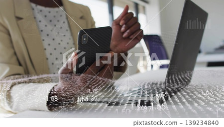 Holding smartphone, woman in blazer gesturing toward data overlays at office, with laptop 139234840
