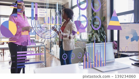 Collaborating women wearing business attire gesturing toward holographic charts in open-plan office Collaborating women wearing business attire gesturing toward holographic charts in open-plan office 139234860