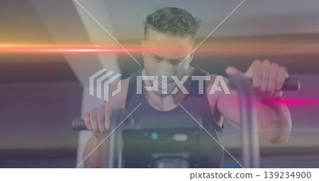 Pedaling male exerciser gripping bike console handlebars in gym, with pink and orange lens flares Pedaling male exerciser gripping bike console handlebars in gym, with pink and orange lens flares 139234900