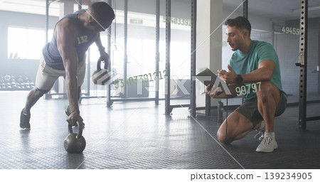 Performing renegade row man holding two kettlebells at gym with trainer tablet, copy space Performing renegade row man holding two kettlebells at gym with trainer tablet, copy space 139234905