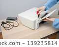 Person performing printer maintenance using a screwdriver to fix a home office printer on a wooden desk, showing hands-on repair, troubleshooting, tools, and small electronics upkeep. 139235814