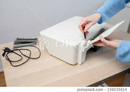 Person performing printer maintenance using a screwdriver to fix a home office printer on a wooden desk, showing hands-on repair, troubleshooting, tools, and small electronics upkeep. Person performing printer maintenance using a screwdriver to fix a home office printer on a wooden desk, showing hands-on repair, troubleshooting, tools, and small electronics upkeep. 139235814