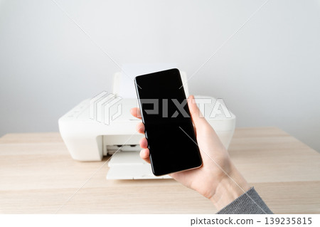 Person holds a smartphone in front of a printer on a wooden desk, wireless mobile printing and connectivity for a modern home office, highlighting convenience, productivity and simple workflow. Person holds a smartphone in front of a printer on a wooden desk, wireless mobile printing and connectivity for a modern home office, highlighting convenience, productivity and simple workflow. 139235815