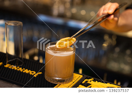 Bartender using precision tweezers to garnish an amber cocktail with a dried orange slice 139237537