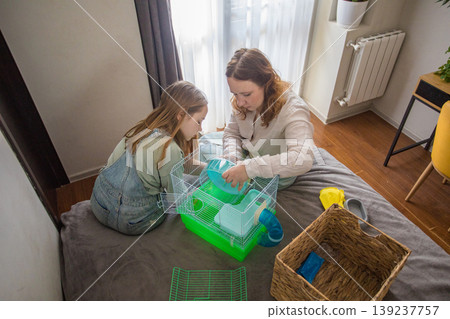 A girl and a woman work together to arrange a green hamster cage on a bed. 139237757
