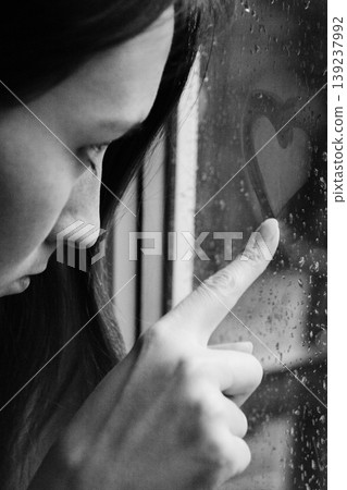 Close-up of a pensive woman drawing a heart on a foggy window 139237992