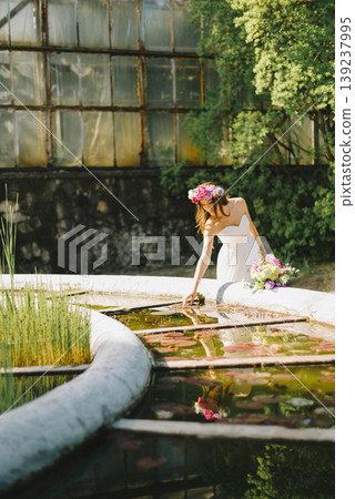 Romantic young girl in light dress with flower wreath sitting near park fountain 139237995
