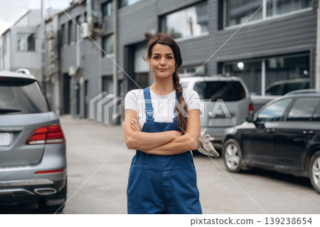 Woman mechanic is standing with arms crossed outdoors against cars and building Woman mechanic is standing with arms crossed outdoors against cars and building 139238654