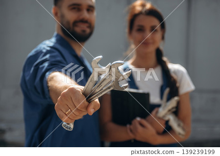Holding tools and digital tablet. Cheerful male and female mechanics are standing outdoors together Holding tools and digital tablet. Cheerful male and female mechanics are standing outdoors together 139239199