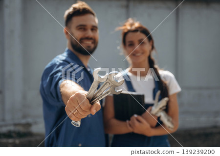 Holding tools and digital tablet. Cheerful male and female mechanics are standing outdoors together 139239200