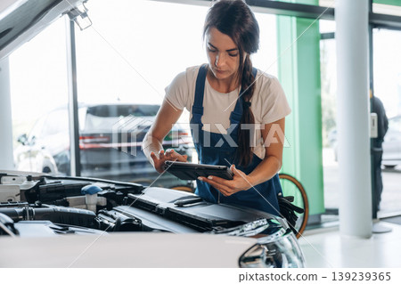 Against window, hood is opened, with digital tablet. Female mechanic is working in the modern car workshop Against window, hood is opened, with digital tablet. Female mechanic is working in the modern car workshop 139239365