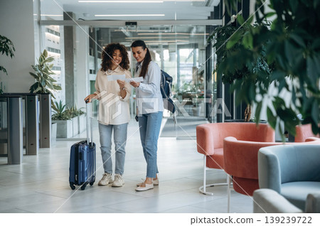 Standing, at daytime. Two women are indoors in the modern hall of building Standing, at daytime. Two women are indoors in the modern hall of building 139239722
