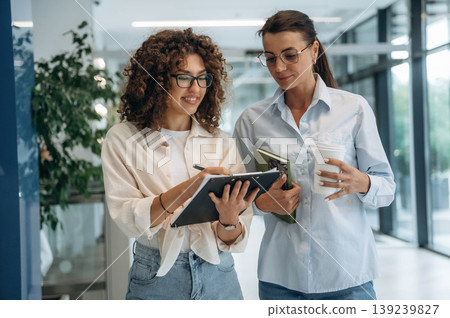 Business partners are together. Two women are indoors in the modern hall of building at daytime Business partners are together. Two women are indoors in the modern hall of building at daytime 139239827