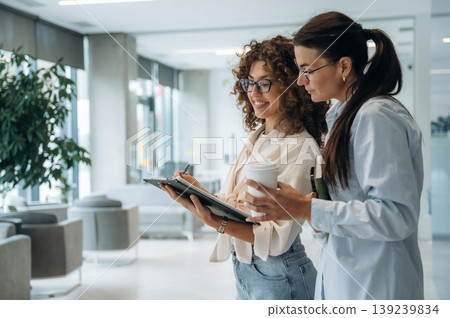 Writing the information into notepad. Two women are indoors in the modern hall of building at daytime Writing the information into notepad. Two women are indoors in the modern hall of building at daytime 139239834