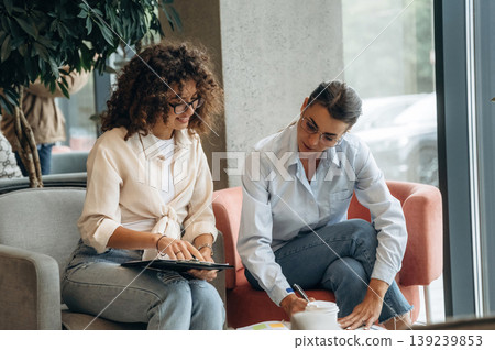 Sitting on the sofa with notepad. Two women are indoors in the modern hall of building at daytime 139239853