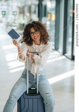 Showing the document, sitting on the suitcase. Beautiful woman is in airport hall Showing the document, sitting on the suitcase. Beautiful woman is in airport hall 139239940