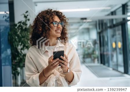 Smartphone is in hands. Pretty woman in casual clothes is in the modern building indoors 139239947