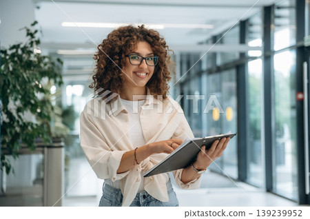 Notepad in hand. Pretty woman in casual clothes is in the modern building indoors Notepad in hand. Pretty woman in casual clothes is in the modern building indoors 139239952