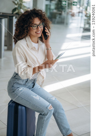 Sitting on the suitcase, with phone in hand. Beautiful woman is in airport hall Sitting on the suitcase, with phone in hand. Beautiful woman is in airport hall 139240061