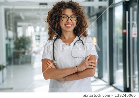 Arms crossed, standing. Woman doctor is standing indoors in the clinic Arms crossed, standing. Woman doctor is standing indoors in the clinic 139240066