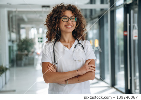 Arms crossed, standing. Woman doctor is standing indoors in the clinic Arms crossed, standing. Woman doctor is standing indoors in the clinic 139240067