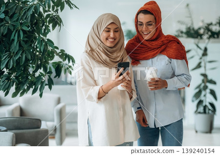 Standing, at daytime. Two women are indoors in the modern hall of building Standing, at daytime. Two women are indoors in the modern hall of building 139240154