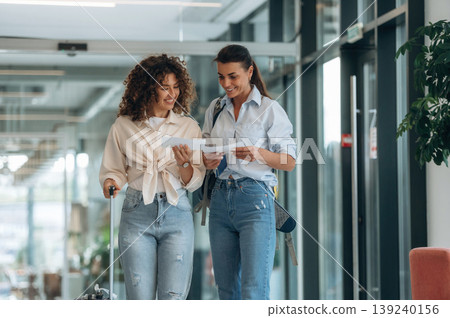 Showing the way, tourist with suitcase. Two women are indoors in the modern hall of building at daytime Showing the way, tourist with suitcase. Two women are indoors in the modern hall of building at daytime 139240156