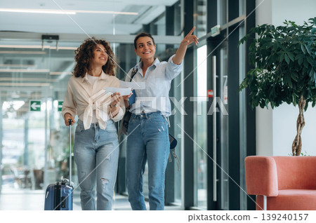 Showing the way, tourist with suitcase. Two women are indoors in the modern hall of building at daytime Showing the way, tourist with suitcase. Two women are indoors in the modern hall of building at daytime 139240157
