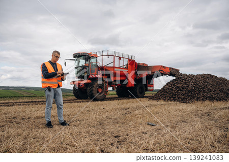 Digital tablet is in hands. Farmer is working in the agricultural field. Against beet harvester Digital tablet is in hands. Farmer is working in the agricultural field. Against beet harvester 139241083