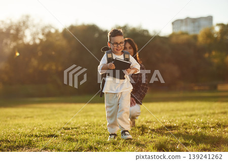 Pupil is with book and backpack. Mother and son together in an open field during daytime Pupil is with book and backpack. Mother and son together in an open field during daytime 139241262